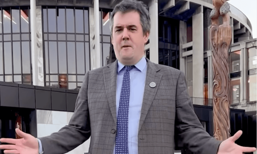 A man (Chris Bishop) in a gray suit and tie stands outdoors with arms open in front of a modern building with large windows and a tall carved wooden post.